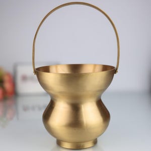 K&ouml;nnte beinhalten: Ein goldfarbener Metallkorb mit gebogenem Griff. Der Korb hat einen abgerundeten Boden und eine breitere Oberseite mit glatter, polierter Oberfl&auml;che. Der Griff ist an den Seiten des Korbes befestigt.