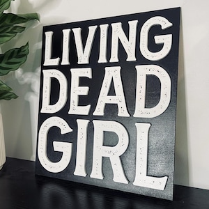 Puede incluir: Un letrero de madera negro con letras blancas que dice "LIVING DEAD GIRL".