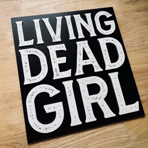 Puede incluir: Un letrero de madera negro con letras blancas que dice "Living Dead Girl".