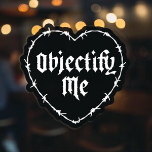 May include: Black and white heart-shaped sticker with barbed wire design. The sticker says "Objectify Me" in a gothic font.