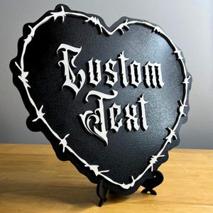 May include: A black heart-shaped sign featuring the words "Custom Text" in a white gothic font. The sign is edged with a white barbed wire design and stands on a black support. Ideal for personalised gifts or home decor.