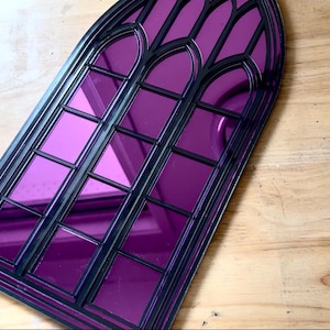 Pode incluir: Um espelho decorativo em forma de janela g&oacute;tica, com moldura preta e vidro espelhado roxo vibrante. O topo arqueado e os pain&eacute;is retangulares criam um efeito visual marcante. O espelho foi concebido para ser montado na parede.
