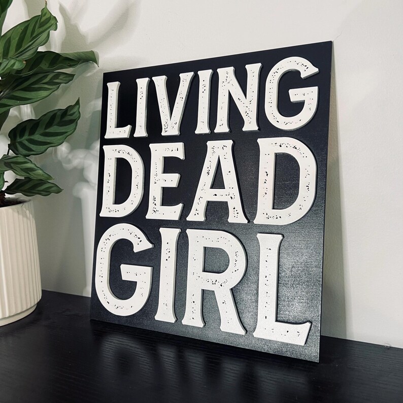 Puede incluir: Un letrero de madera negro con letras blancas que dice "LIVING DEAD GIRL".