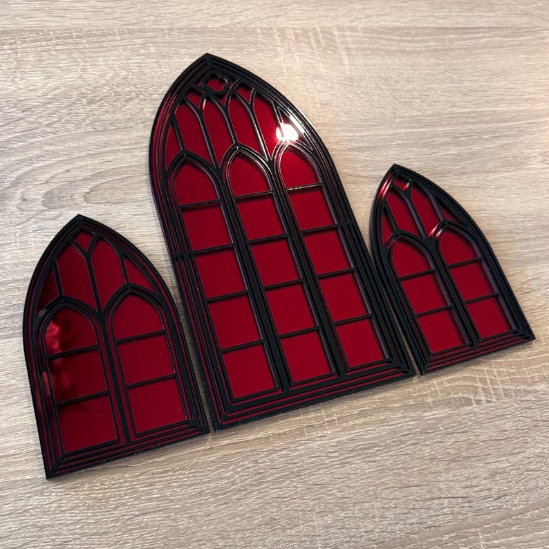 Pode incluir: Tr&ecirc;s decora&ccedil;&otilde;es de janelas em estilo g&oacute;tico. Cada janela apresenta uma superf&iacute;cie espelhada vermelha com uma moldura preta. As janelas est&atilde;o dispostas em um layout escalonado, com a maior no centro e duas menores em cada lado.