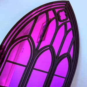 Pode incluir: Um espelho de estilo g&oacute;tico com uma superf&iacute;cie reflexiva rosa vibrante. O espelho &eacute; emoldurado por uma borda escura e ornamentada, com pain&eacute;is arqueados em forma de janela e detalhes decorativos. A forma geral lembra uma janela de catedral.