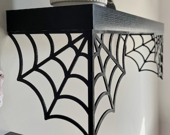 Puede incluir: Estante de madera negro con soportes de telaraña. El estante está montado en una pared blanca y tiene una parte superior negra. Una maceta de cerámica blanca se encuentra en el estante.