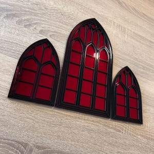 Pode incluir: Tr&ecirc;s decora&ccedil;&otilde;es de janelas em estilo g&oacute;tico com moldura preta e vidro vermelho. As janelas s&atilde;o de tamanhos variados, sendo a maior ao centro. O design lembra a arquitetura das catedrais.