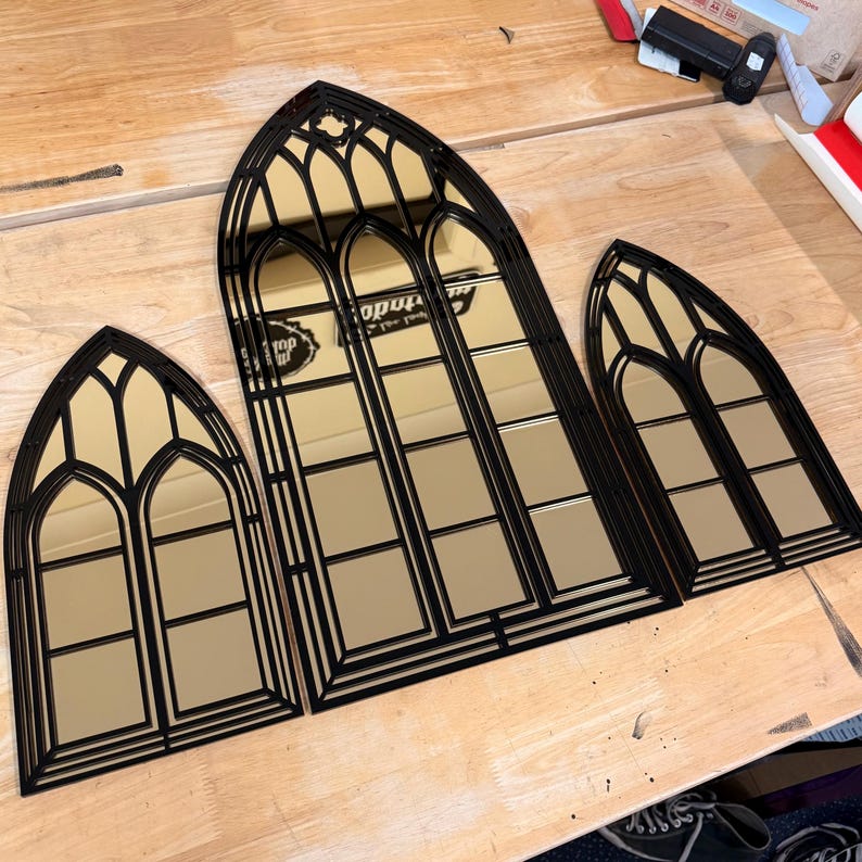 Pode incluir: Tr&ecirc;s espelhos pretos em forma de janela g&oacute;tica com superf&iacute;cies refletoras douradas. Os espelhos est&atilde;o dispostos sobre uma superf&iacute;cie de madeira, mostrando seu intrincado design arquitet&ocirc;nico. Os espelhos variam em tamanho.