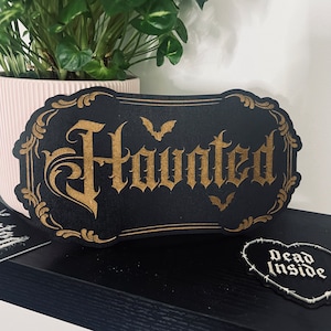 Puede incluir: Un letrero de madera negro con letras doradas que dice "Haunted". El letrero tiene un borde decorativo y siluetas de murciélagos.