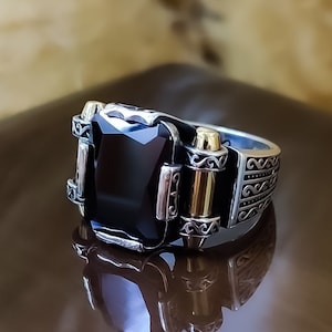 May include: A silver ring featuring a large, rectangular, faceted black stone set in the center. The band is decorated with intricate scrollwork patterns and includes two vertical gold-toned accents flanking the central stone on each side.