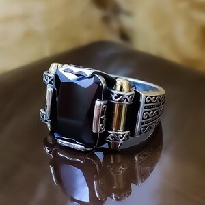 May include: A silver-toned ring featuring a large, rectangular, faceted black stone set in the center. The band is decorated with intricate scrollwork patterns and includes two vertical gold-toned accents flanking the central black stone.