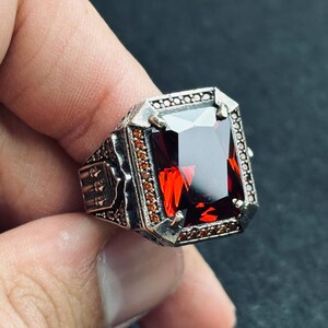 Op de afbeelding: Een zilveren ring met een grote, rechthoekige, dieprode edelsteen. De ring heeft ingewikkelde details op de band en rond de steen. De zetting is zilver met kleine, roodbruine accentstenen. De ring wordt tegen een donkere achtergrond gehouden.