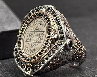 May include: A silver ring featuring an oval center plate with an engraved Star of David and Arabic script, surrounded by a border of small black stones. The band is decorated with intricate metalwork and additional black stone accents.