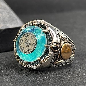 May include: A silver ring featuring a large blue stone set in a silver and gold setting. The stone displays a six-pointed star design with Arabic script.