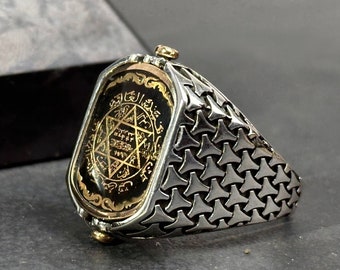 May include: A silver-toned ring featuring a rectangular face with a black and gold six-pointed star emblem and Arabic script. The band has a textured, repeating triangular pattern and small gold-colored accents at the top and bottom.