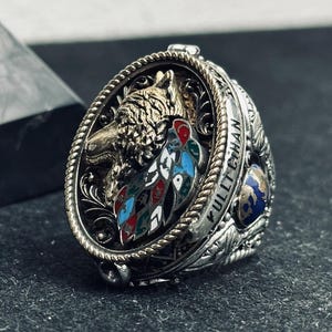 May include: Silver-toned ring with an oval centerpiece featuring a detailed wolf head and colorful feather design. The ring has intricate side detailing and the word "KULLTCHAN" engraved on the band. The ring is set against a dark background.