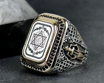 May include: A silver ring with a black and white rectangular design featuring a six-pointed star and Hebrew text. The ring has a gold trim and a detailed design on the band.
