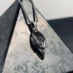 May include: A silver-toned pendant necklace featuring a detailed raven's head design. The raven's head is adorned with black gemstones and hangs from a matching chain. The necklace is displayed on a textured, gray surface.