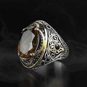 Puede incluir: Un anillo de plata con una gran piedra preciosa ovalada de color ámbar. El anillo presenta intrincados detalles de filigrana negra y dorada alrededor de la piedra preciosa y en la banda. Pequeñas piedras negras acentúan el diseño.