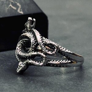 Op de afbeelding: Een zilverkleurige metalen ring met een ontwerp van een kronkelende slang die om de vinger wikkelt. De slang heeft gedetailleerde schubben en een duidelijke kop aan elk uiteinde van de winding, wat een gestructureerde ring vormt.