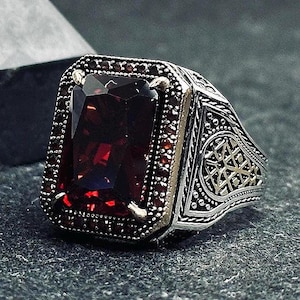 May include: A silver ring featuring a large, rectangular, deep red gemstone. The ring is adorned with smaller red stones around the main stone and detailed patterns on the sides, including a gold-coloured star design.