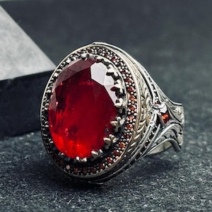 May include: A silver ring featuring a large, oval-cut, deep red gemstone. The ring is embellished with smaller red stones and detailed metalwork around the central stone and the band. The design of the ring is classic and elegant, with intricate detailing.