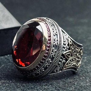 May include: A silver ring with an oval, faceted, deep red gemstone. The ring has a gold-colored band around the gemstone and is accented with small red stones. The band features intricate, ornate detailing.