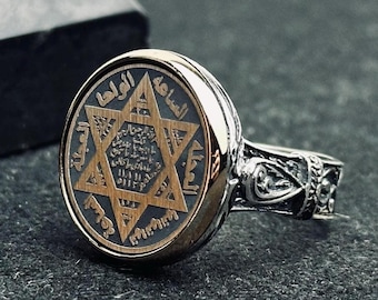 May include: A silver ring with an oval face featuring a gold Star of David design on a dark background. The ring has intricate detailing on the band and the face has Arabic script around the star. The ring is set against a dark background.