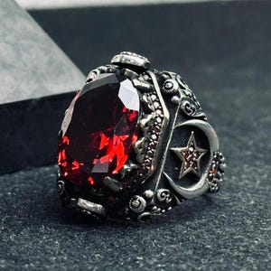 May include: A silver ring with a large, oval, deep red gemstone as the centerpiece. The ring features intricate detailing, including a crescent moon and star design on the side, and smaller red stones.
