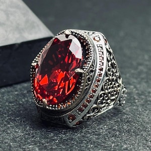 May include: A silver ring with an oval, faceted, deep red gemstone. The ring has an ornate, filigree design on the band and around the gemstone. Small red stones accent the band. The ring is a statement piece.