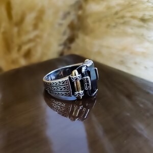 May include: A silver ring featuring an elongated rectangular black stone set in a metallic frame. The band displays intricate engraved scrollwork patterns along the sides, accented by a small gold-toned cylindrical detail near the central stone.