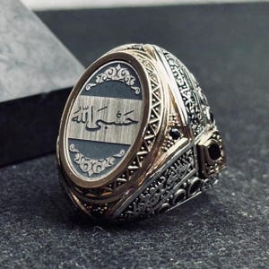May include: A silver and gold-toned ring featuring an oval face with a brushed metal center plate inscribed with Arabic calligraphy. The band and setting are decorated with intricate engraved patterns and small black stone accents on the sides.
