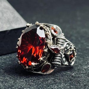 May include: A silver ring with a large, oval-cut, deep red gemstone. The ring has an ornate design with smaller red accent stones and detailed metalwork. The ring's design is intricate.