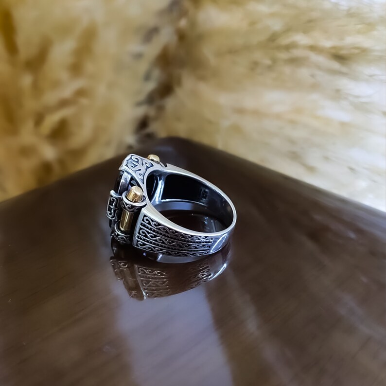 May include: A silver ring featuring intricate engraved patterns along the band and a central dark stone setting. The design includes two small, polished gold-toned cylindrical accents positioned vertically on the sides of the main setting.