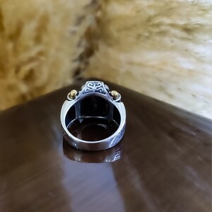 May include: A silver ring featuring a large, dark rectangular stone set in the center. The band displays intricate engraved patterns on the sides, accented by two small, polished gold-tone spheres positioned on either side of the stone setting.