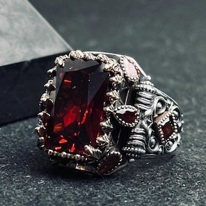 以下が含まれることがあります： 大きな長方形の深い赤色の宝石を特徴とするシルバーリング。リングは、小さな赤いアクセントストーンと詳細なバンドを備えた、複雑で装飾的なディテールを備えています。デザインはヴィンテージにインスパイアされています。