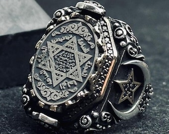 May include: A silver-toned ring featuring an oval face with an engraved six-pointed star and Arabic script. The sides of the band are decorated with intricate scrollwork, a crescent moon, and a small star set with tiny black stones.