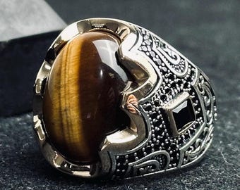 May include: A silver ring featuring a large, oval, brown tiger's eye gemstone at the center. The band is decorated with intricate metal engravings, small black stones, and a single square black stone set on the side of the ring.