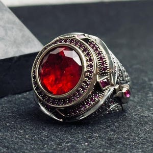 May include: A silver ring with a large, round, red gemstone surrounded by smaller, purple gemstones. The ring has intricate details and a textured band.