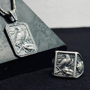 Op de afbeelding: Een zilveren hangerketting en ring set. De rechthoekige hanger en ring hebben een gedetailleerd reliëf van een adelaar boven een wolf. De sieraden zijn gemaakt van een glanzend, metalen materiaal.
