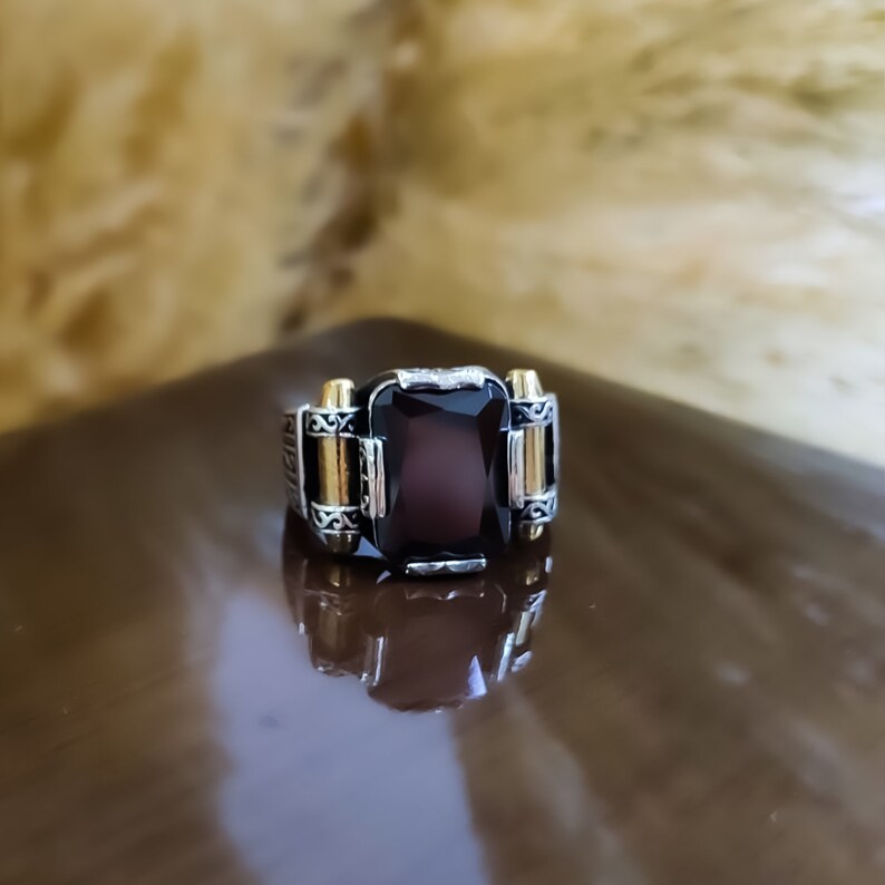 May include: A silver-toned ring featuring a large, rectangular, dark brown faceted gemstone set in the center. The band is accented with gold-toned cylindrical side details and intricate engraved scrollwork patterns on the metal surface.