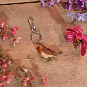 May include: A keychain featuring a small, brown bird figurine with a distinct orange-red breast and a cream-colored belly. The bird is attached to a silver-toned metal split ring and a lobster claw clasp, providing a secure way to attach keys.