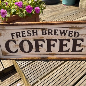 May include: A rectangular wooden sign with a distressed white background and a dark brown wooden frame. The sign features the text "FRESH BREWED COFFEE" in bold, dark brown, serif lettering, with visible weathered details across the surface.