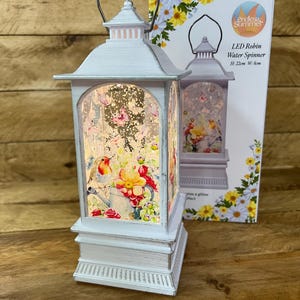 May include: A white LED lantern with a bird and floral design. The lantern has a handle and a decorative base. The box next to the lantern reads "LED Robin Water Spinner" and the dimensions are H: 22cm W: 8cm.