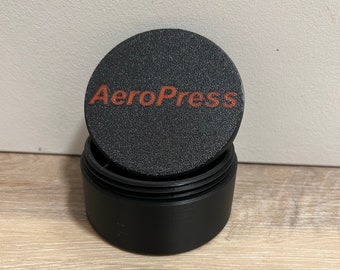 Könnte beinhalten: Eine schwarze AeroPress Kaffeemaschine mit einem strukturierten Deckel. Der Deckel trägt den Schriftzug "AeroPress" in orange.