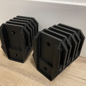 Könnte beinhalten: Zwei schwarze, 3D-gedruckte Objekte im Ziehharmonika-Stil. Jedes Objekt hat eine Reihe rechteckiger Paneele, die in einem Zickzackmuster verbunden sind. Die Objekte haben ein geometrisches Design mit Löchern zur Befestigung. Sie liegen auf einer hellen Holzoberfläche.