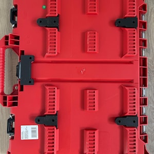 May include: A rectangular red plastic tool storage case featuring a moulded exterior with multiple raised ribs, black locking latches, and a side handle. A white rectangular product label with black text and a barcode is affixed to the lower left corner.