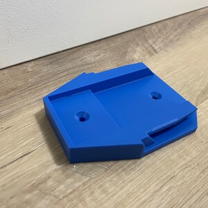 Może przedstawiać: Jasnoniebieski, geometryczny plastikowy przedmiot z dwoma okrągłymi otworami i zagłębionym obszarem. Przedmiot spoczywa na jasnej drewnianej powierzchni. Obiekt wydaje się być wspornikiem montażowym lub uchwytem.