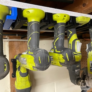 Peut inclure: Plusieurs outils électriques, dont des perceuses et une meuleuse, suspendus à un support de rangement noir et bleu. Les outils sont principalement noirs et jaunes, avec des marques de fabricants visibles. L'ensemble suggère un atelier ou un garage.