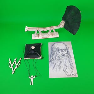 May include: A wooden model of a parachute with a black fabric canopy, a small paper figure hanging from the parachute, and a pair of black and silver compasses. The model is on a green background. There is also a drawing of Leonardo da Vinci on a white piece of paper with the text "Studio Box" in the bottom right corner.